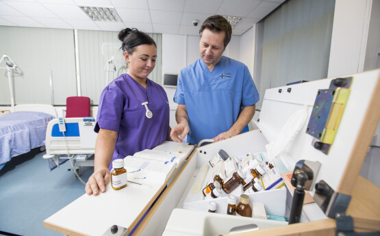 Nursing student wearing purple scrubs, and a lecturer wearing blue scrubs. They are working together and looking at different medicines. 