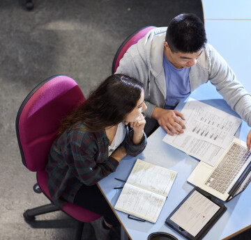 Two students working together at a library desk with a laptop, papers and notebooks