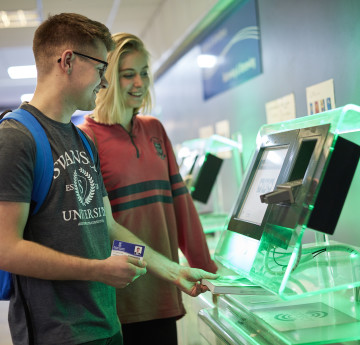 two students using the self service terminals in the library to borrow books