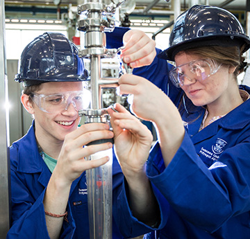 Two students in blue protective suits and wearing hart hats examine mechanical equipment