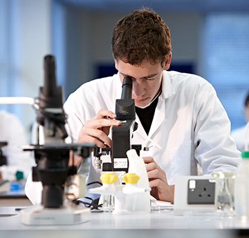 A male student in white lab coat looking through a microscope
