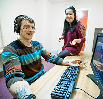 Student wearing electrodes on his head using a computer, with another person sat on the desk instructing him