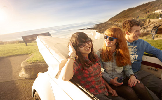 Three students sitting in the back of a white convertible car with Bracelet Bay in the background
