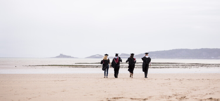 Four students walking on a beach in graduation gowns 