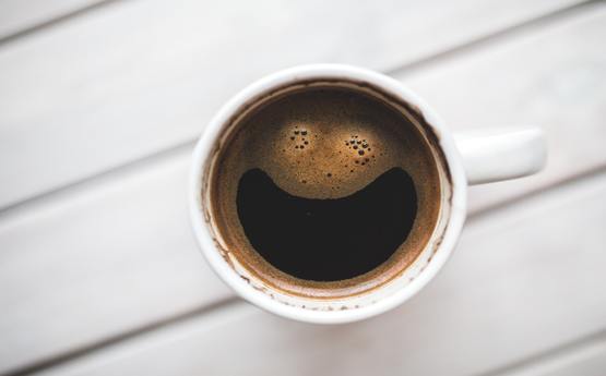 A mug of coffee placed on a white, wooden desk. The foam of the coffee in the shape of a smiling face.