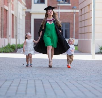 Young woman in academic dress holding hands with two young children walking through Swansea University Bay Campus