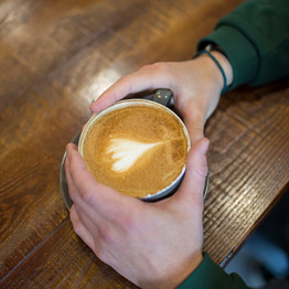 A person's hands resting on a table holding a cup of coffee with a heart shape in the foam